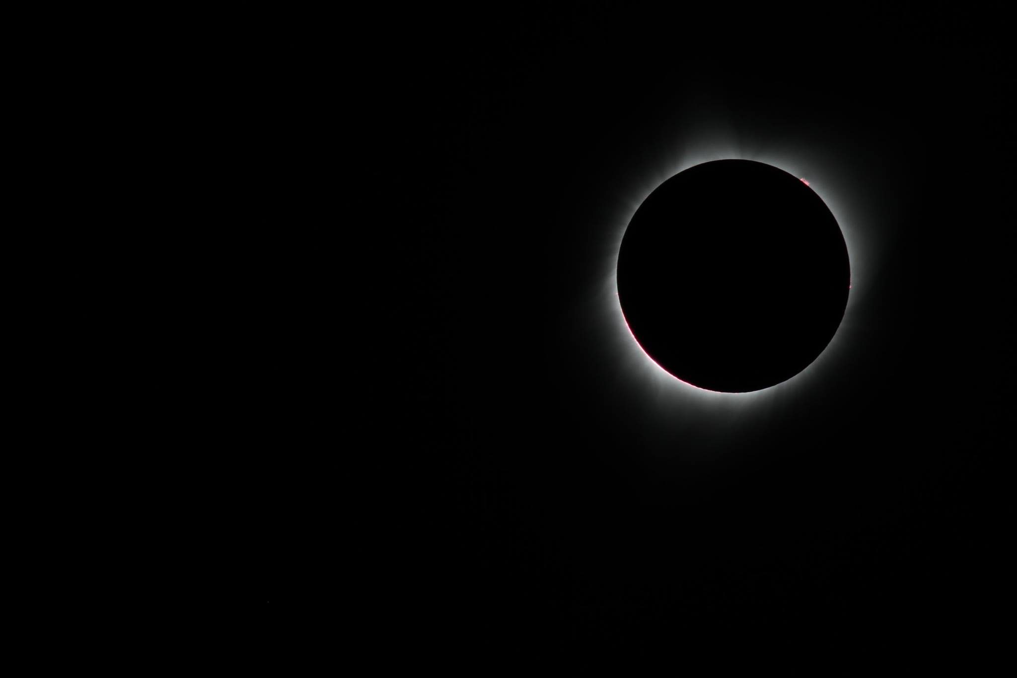 a solar eclipse where the moon has completely blotted out the sun to black except for a faint white glow at the edge and a small reddish-pink burst at the top right